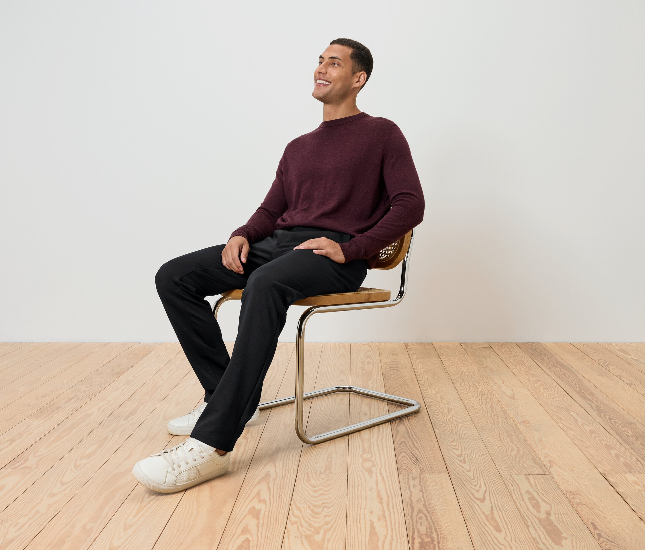 Egy férfi ül egy széken bordó pulóverben, fekete nadrágban és fehér cipőben, és felfelé néz.