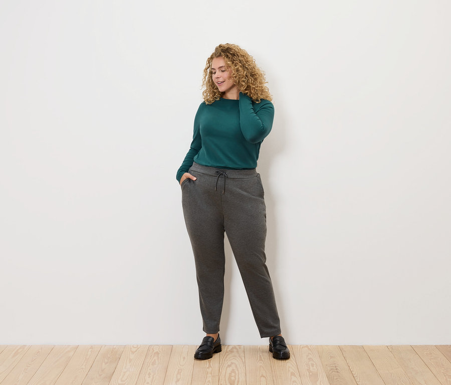 Egy göndör hajú nő pózol világos háttér előtt. Zöld, hosszú ujjú blúzt és szürke joggingalsót visel.