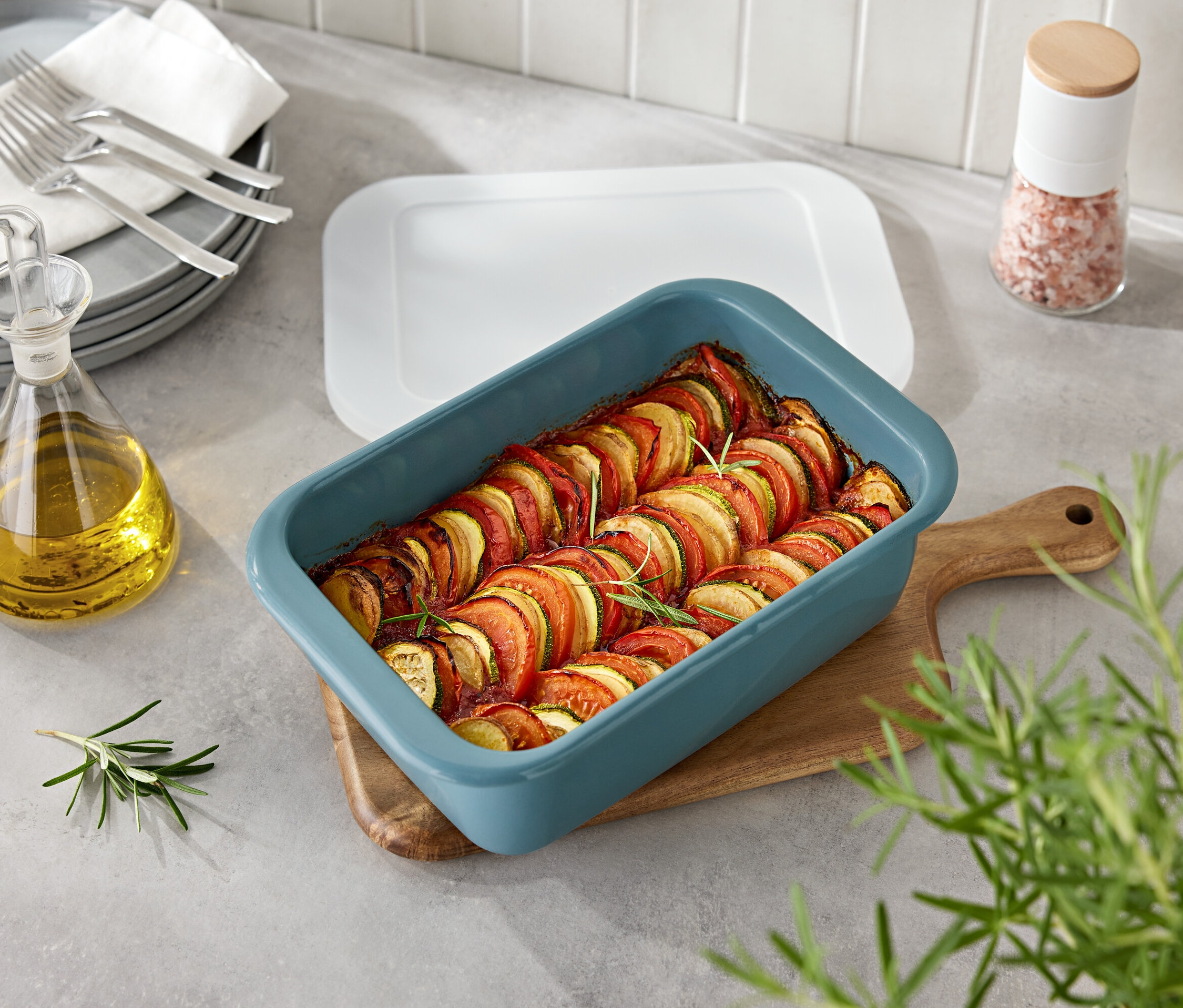 Zöldségek kék sütőformában, fa deszkán. Mellette egy üveg olaj és egy fűszertartó.