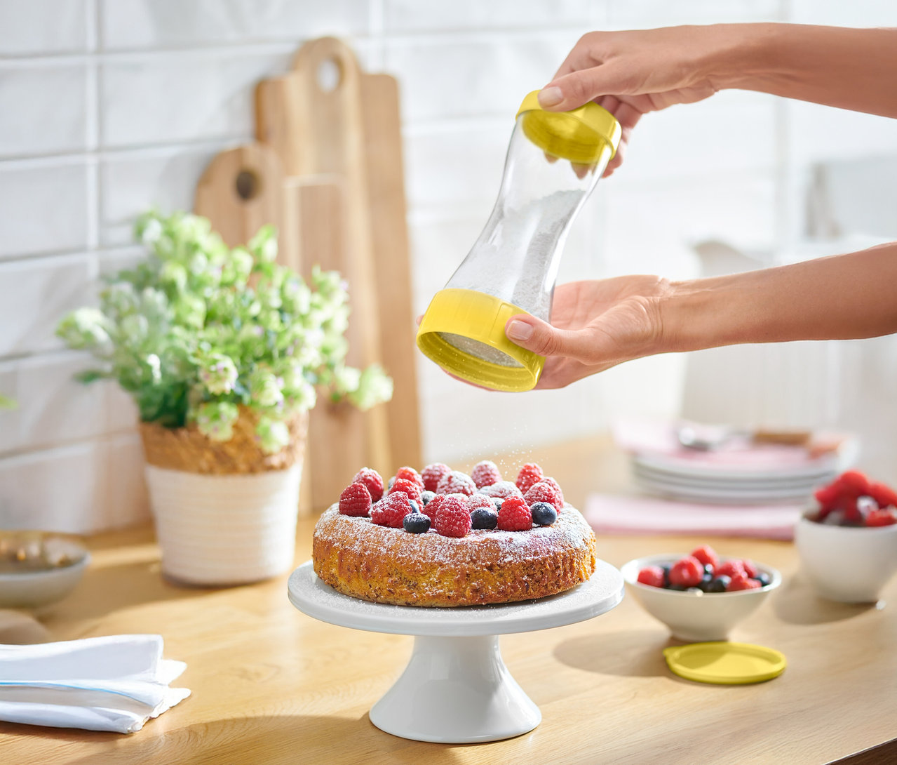 Egy személy porcukrot szór egy bogyós gyümölcsös tortára egy sárga szórófejjel.