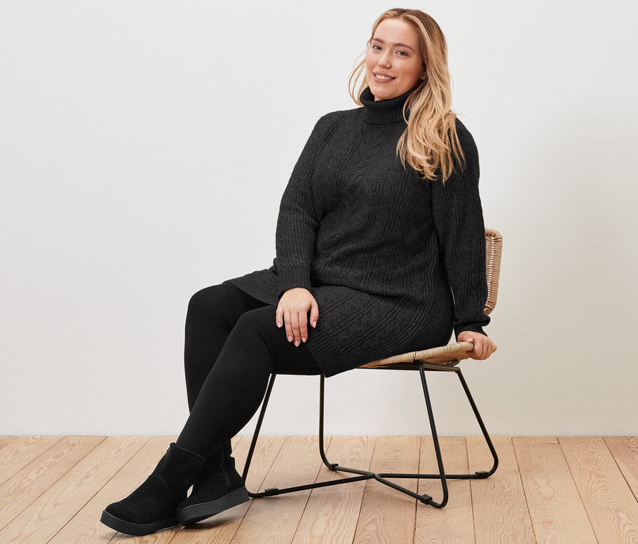 Egy nő ül egy széken, és egy hosszú, fekete, fonott mintás kötött pulóvert visel.