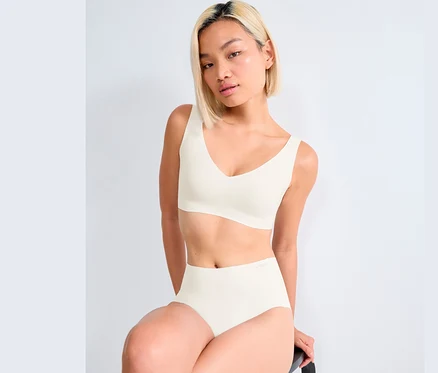 Egy nő ül, és fehér sloggi ZERO Feel 2.0 bralette-et és alsóneműt visel.
