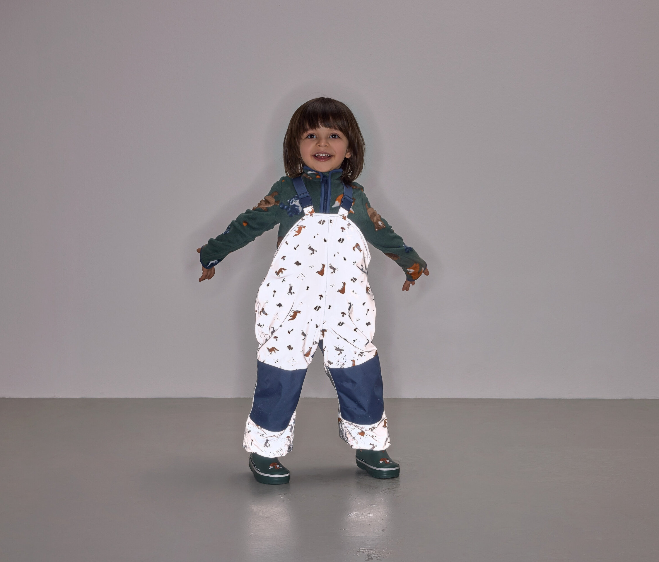 Egy mosolygó gyermek áll széttárt karokkal, állatmintás kantáros nadrágot és sötétkék nadrágszárat visel.