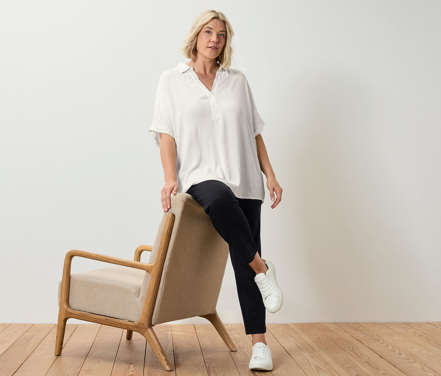 Egy szőke nő egy bézs színű fotel mellett pózol, krémfehér blúzt visel.