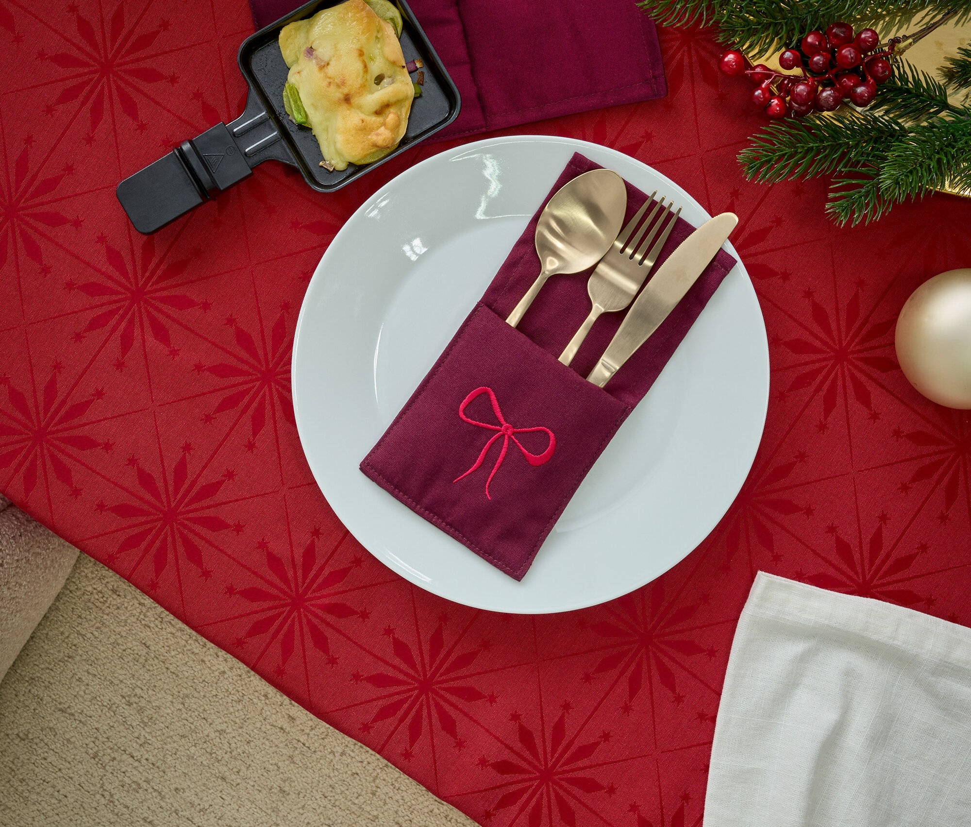 Tányér hímzett szalvétatartóval, arany evőeszközökkel, raclette serpenyővel és karácsonyi dekorációval, piros asztalterítőn.