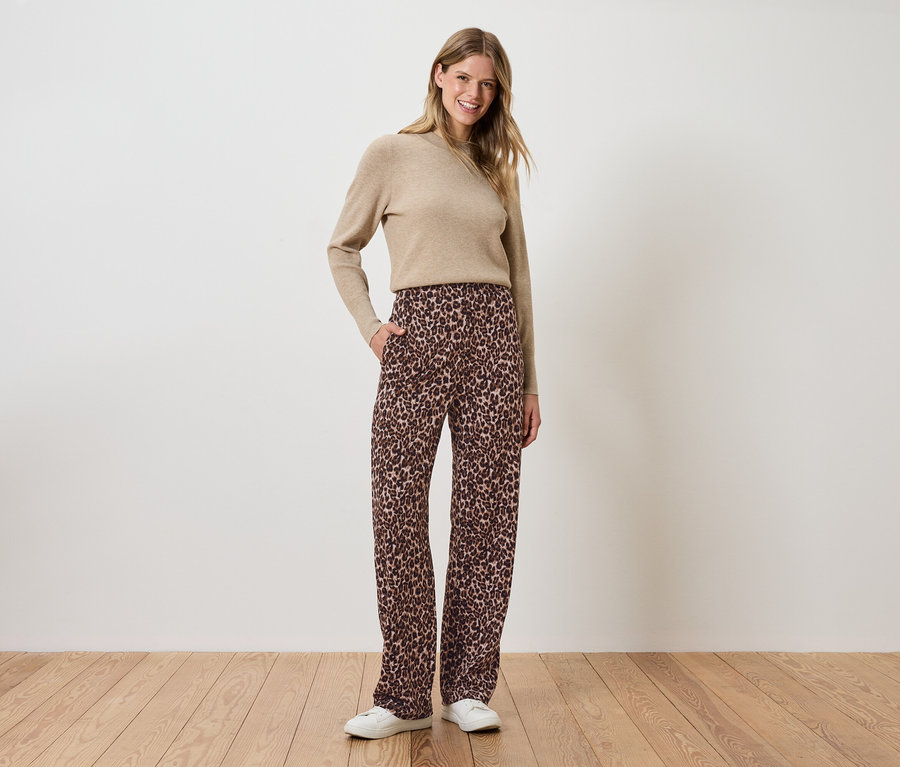 Egy nő áll, kezét a barna, virágos nadrágjának zsebébe dugva, és egy bézs színű pulóvert visel.