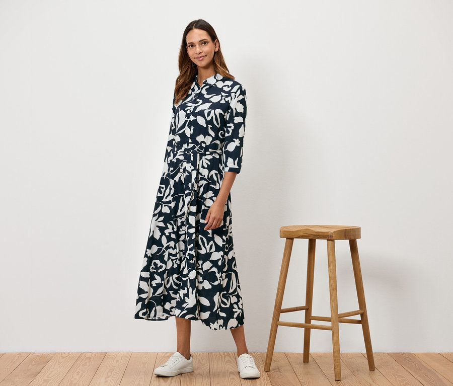 Egy nő áll egy fából készült zsámoly mellett, és egy sötétkék, fehér virágmintás ruhát visel.