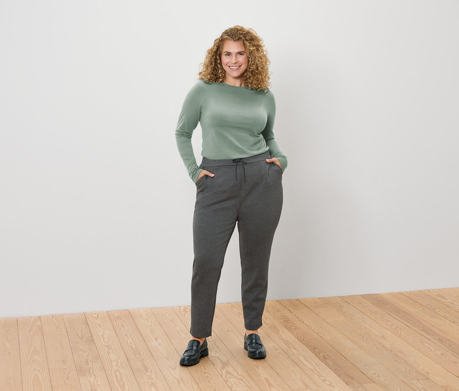 A göndör szőke hajú nő zöld, hosszú ujjú inget és szürke nadrágot visel, kezét a zsebében tartja.