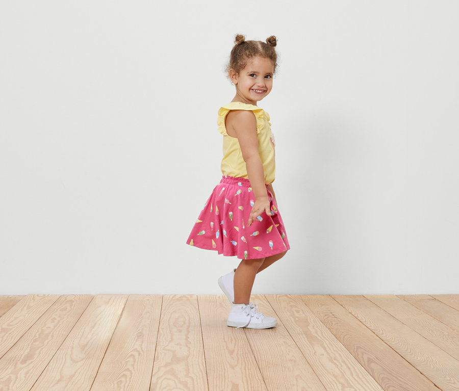 Mosolygó kislány oldalra pózol egy kislány jersey szoknyában, fagyi mintás nadrággal és egy sárga pólóban.