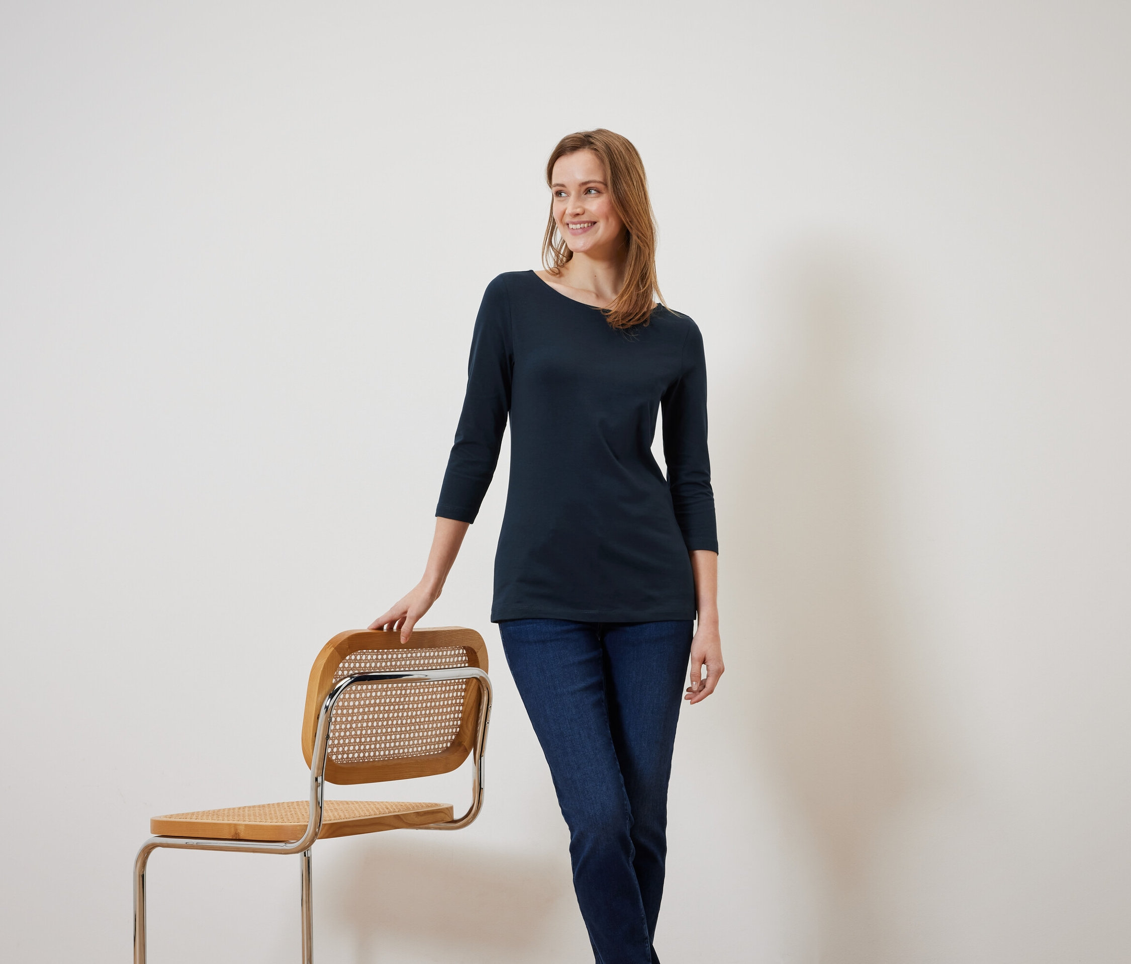Egy mosolygó nő háromnegyedes ujjú, sötétkék blúzban egy fa széknek támaszkodik.