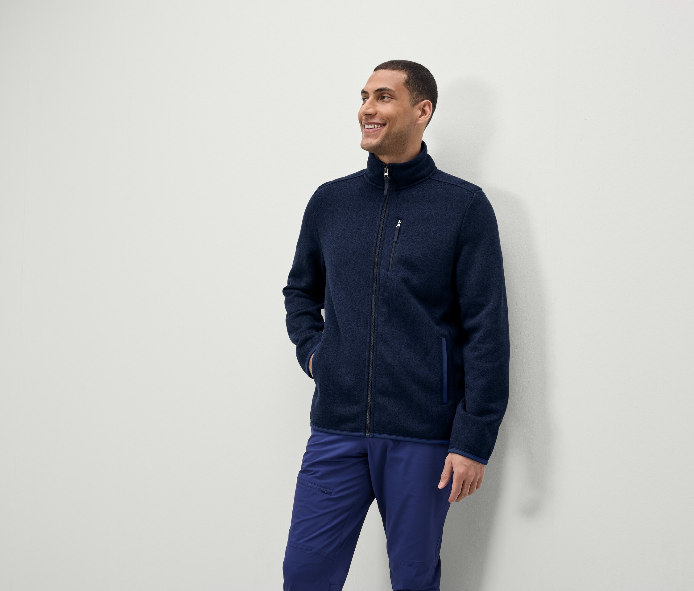Egy férfi mosolyog és oldalra néz, kék cipzáros fleece kabátot és kék nadrágot visel.