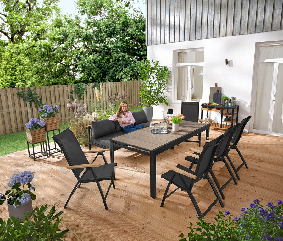 Egy nő ül egy 2 az 1-ben kerti padon ‒ »Elin« napozóágyon. A háttérben »Elin« 2 virágtartó láda áll. A teraszon van még egy grillkocsi policement munkalappal és egy kihúzható kerti asztal Duraboard asztallappal.