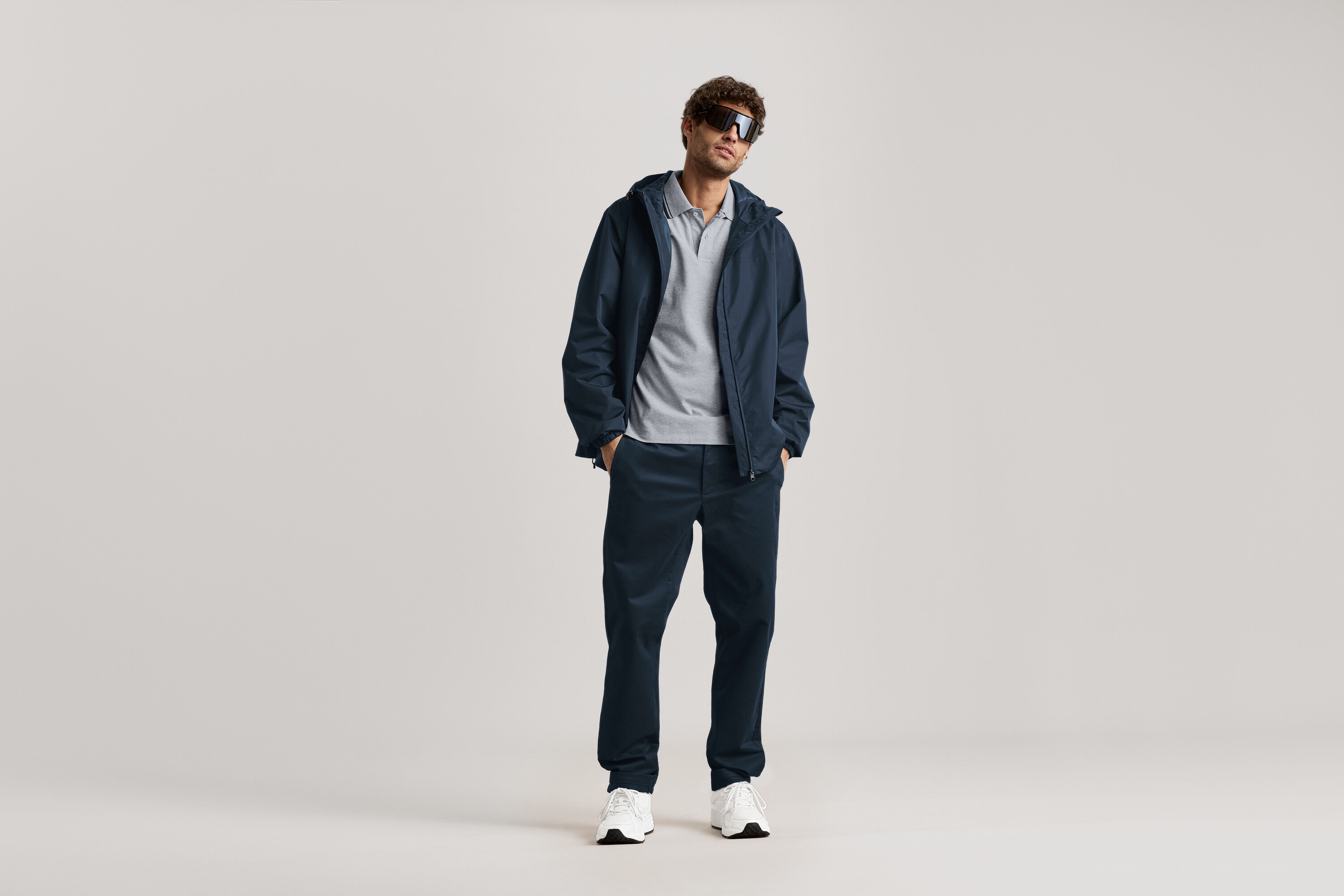 Egy férfi áll napszemüvegben, sötétkék kabátban és sötétkék chino nadrágban.