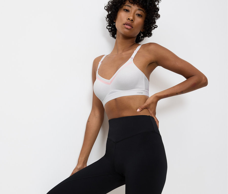 Nő pózol fehér sportmelltartóban rózsaszín részletekkel és fekete leggingsben, fehér fal előtt.