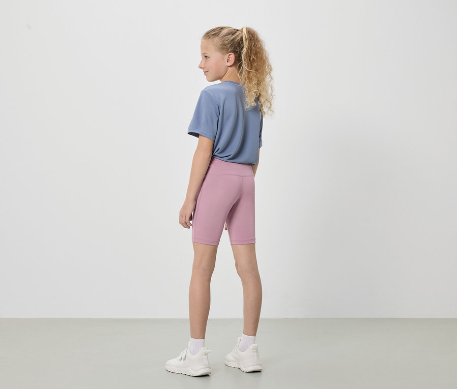 A szőke hajú lány hátulról látható, kék pólót, rövid, rózsaszín leggingset és fehér tornacipőt visel fehér háttér előtt.
