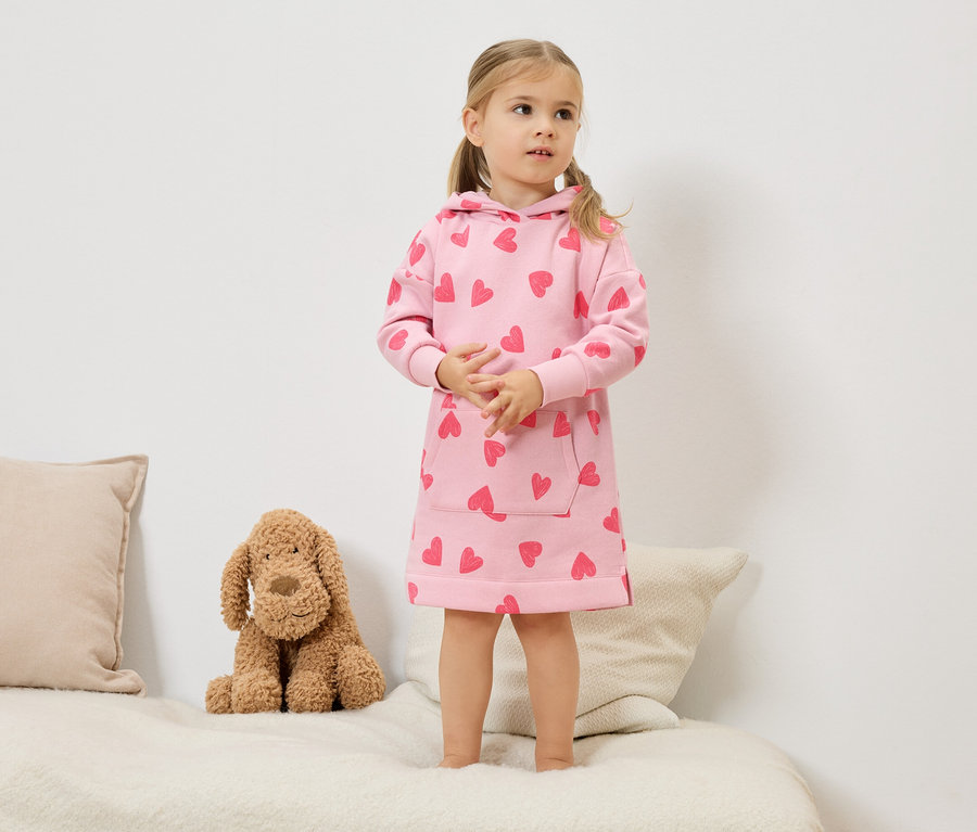 Egy szőke, copfos kislány áll az ágyon, rózsaszín, szívecskés kapucnis ruhában. Mellette egy plüsskutya ül.