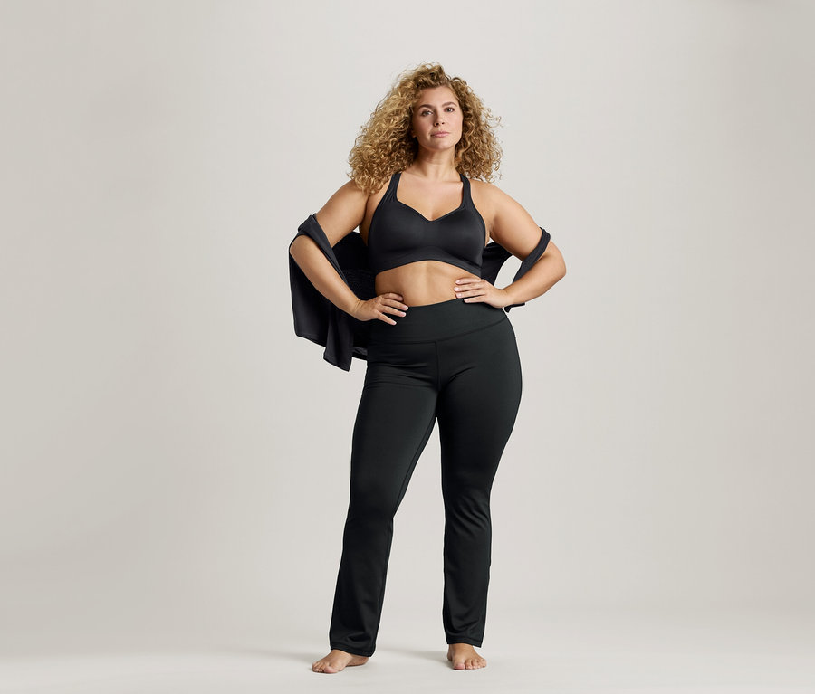 Göndör hajú nő fekete sportmelltartóban és leggingsben, kabáttal a vállán és csípőn tartott kézzel.