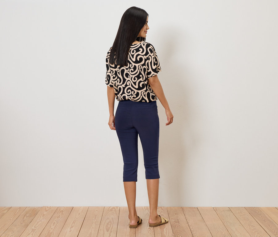 Hátulról látható nő mintás felsőben, kék capri nadrágban és szandálban, fehér fal előtt.