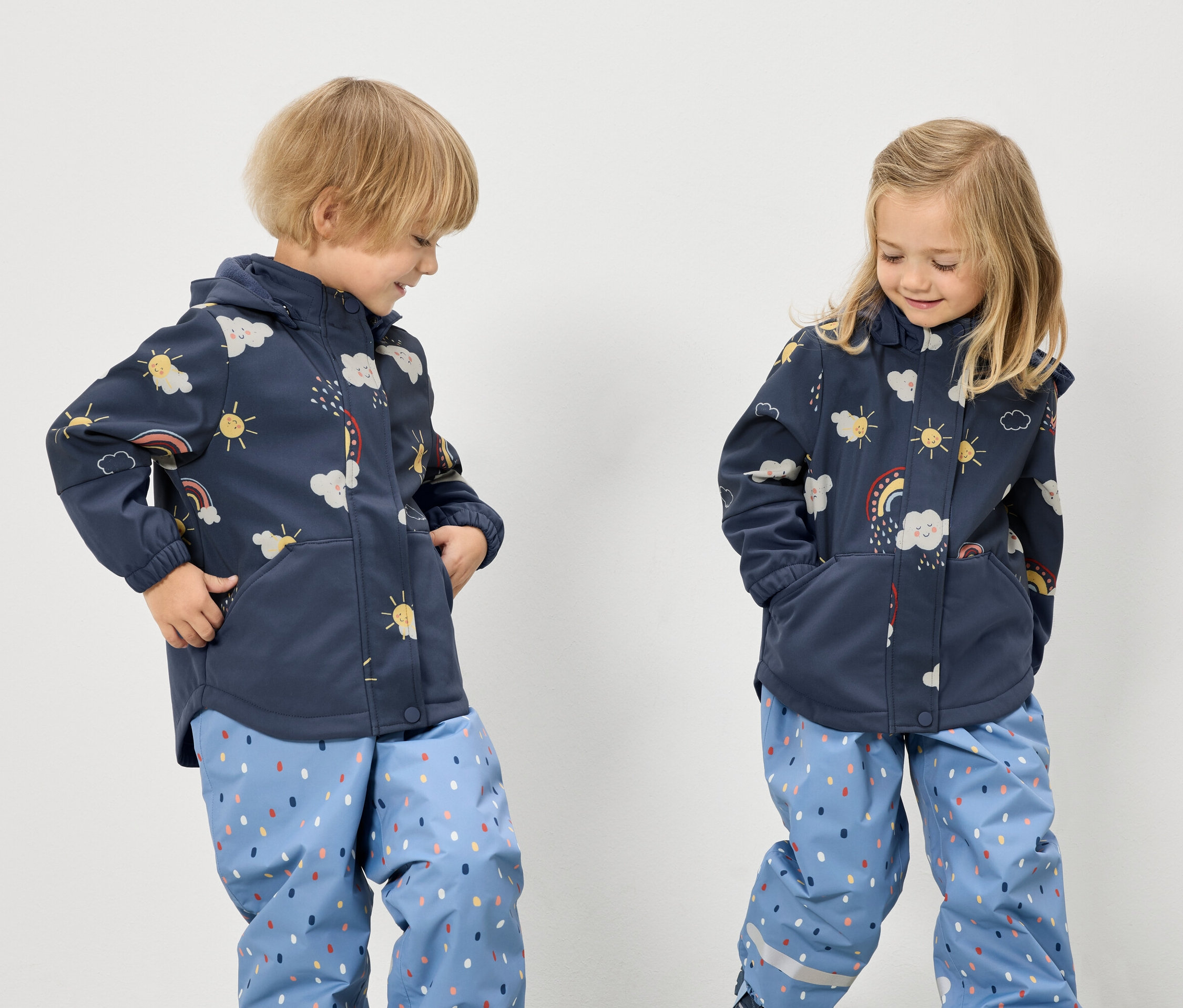 Két gyerek áll kék, szivárványos, felhős és napmintás kabátban és pöttyös kék nadrágban, fehér háttér előtt.
