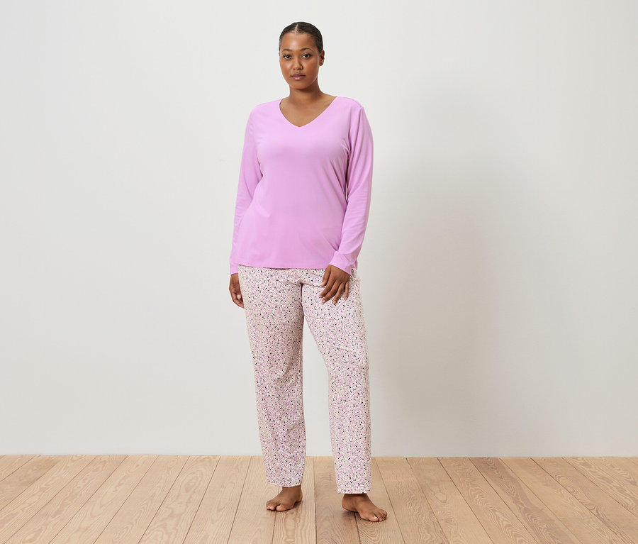 Egy nő áll mezítláb egy fehér fal előtt, lila pizsamafelsőt és rózsaszín, virágmintás pizsamanadrágot visel.