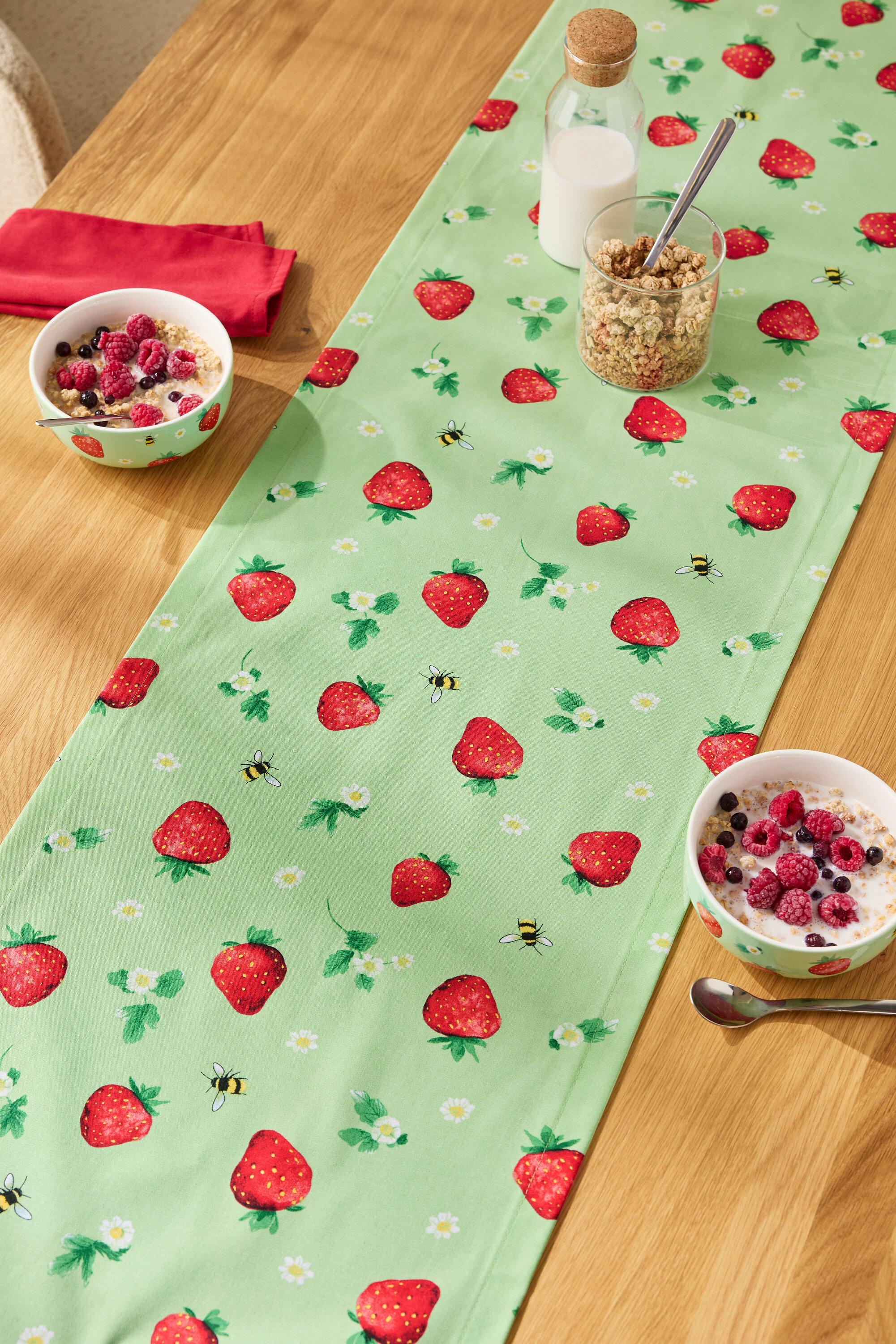 Egy fa asztalon egy epermintás asztali futó, rajta müzlis tálak, egy pohár müzli és egy üveg tej.