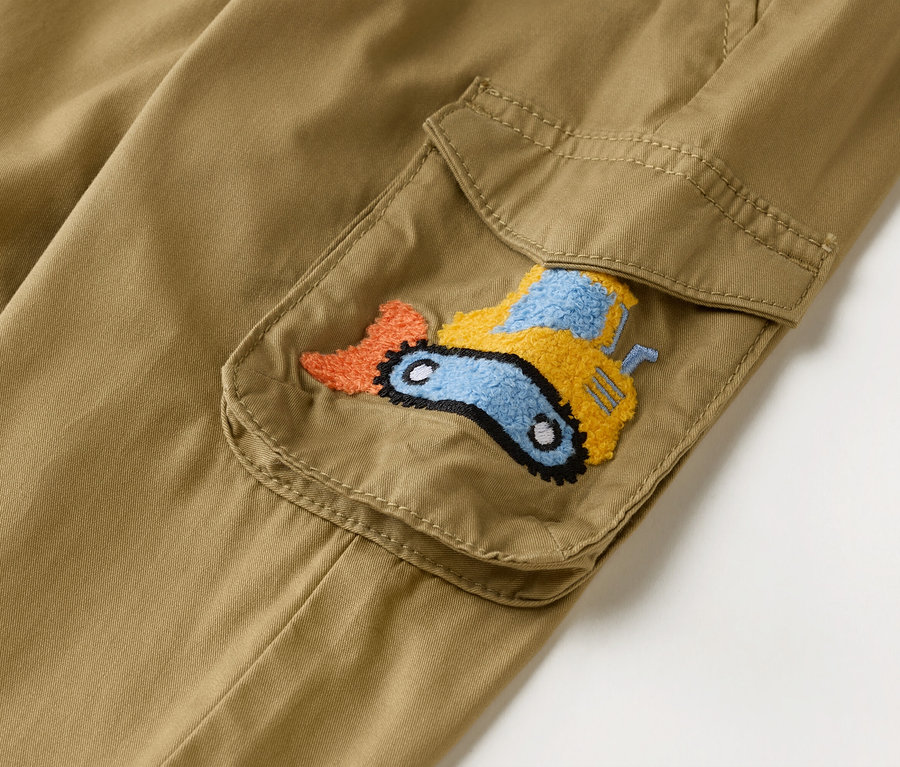 Egy khaki színű cargo nadrág közeli képe, rátétes zsebbel, amelyet narancssárga, sárga és kék hímzett kotrógép díszít.