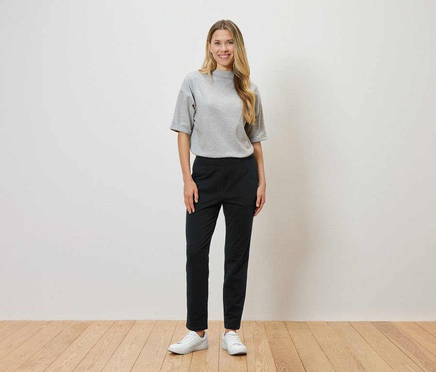 Egy szőke hajú nő áll egy fehér fal előtt, szürke pólót, fekete nadrágot és fehér tornacipőt visel.