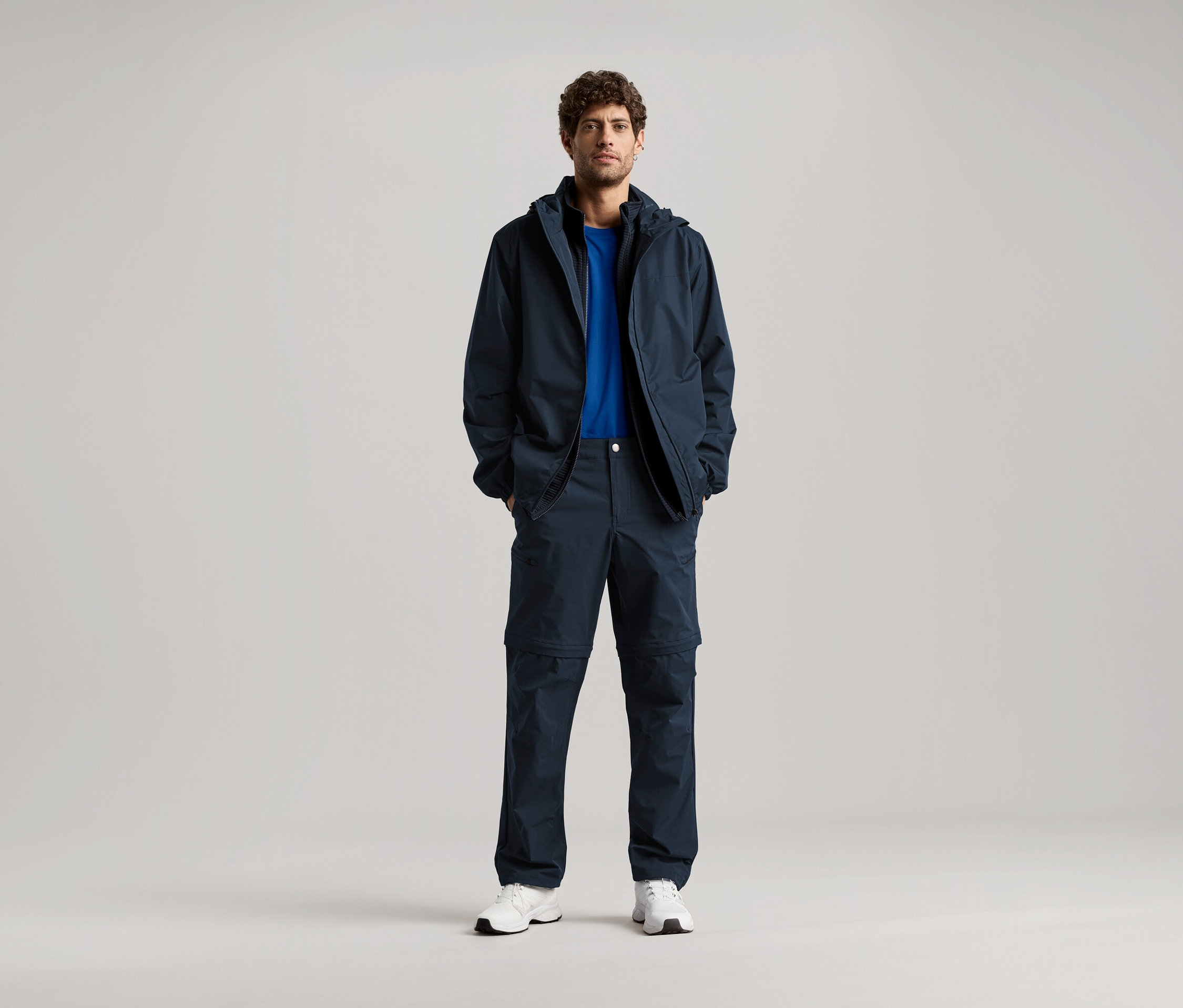 Egy férfi áll sötétkék kabátban és hozzáillő nadrágban, kezét a zsebében tartja, kék pólót és fehér tornacipőt visel.