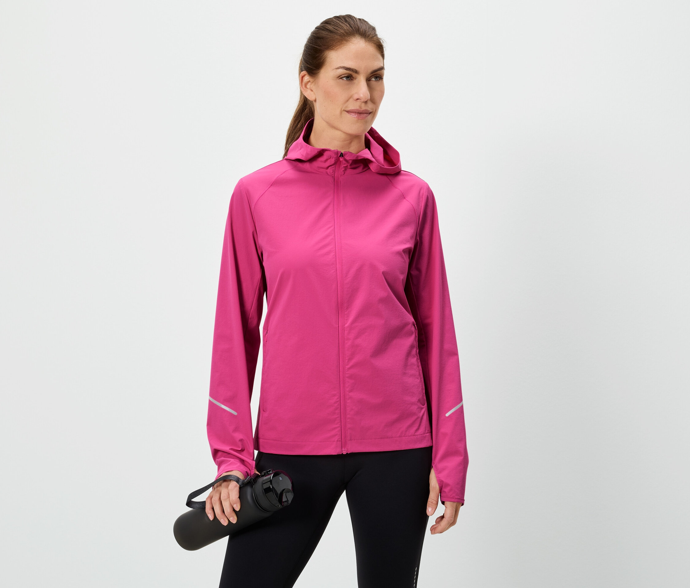 Nő tart egy fekete vizes palackot, rózsaszín kabátot és fekete leggingset visel fehér háttér előtt.