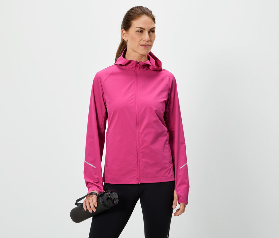 Nő tart egy fekete vizes palackot, rózsaszín kabátot és fekete leggingset visel fehér háttér előtt.