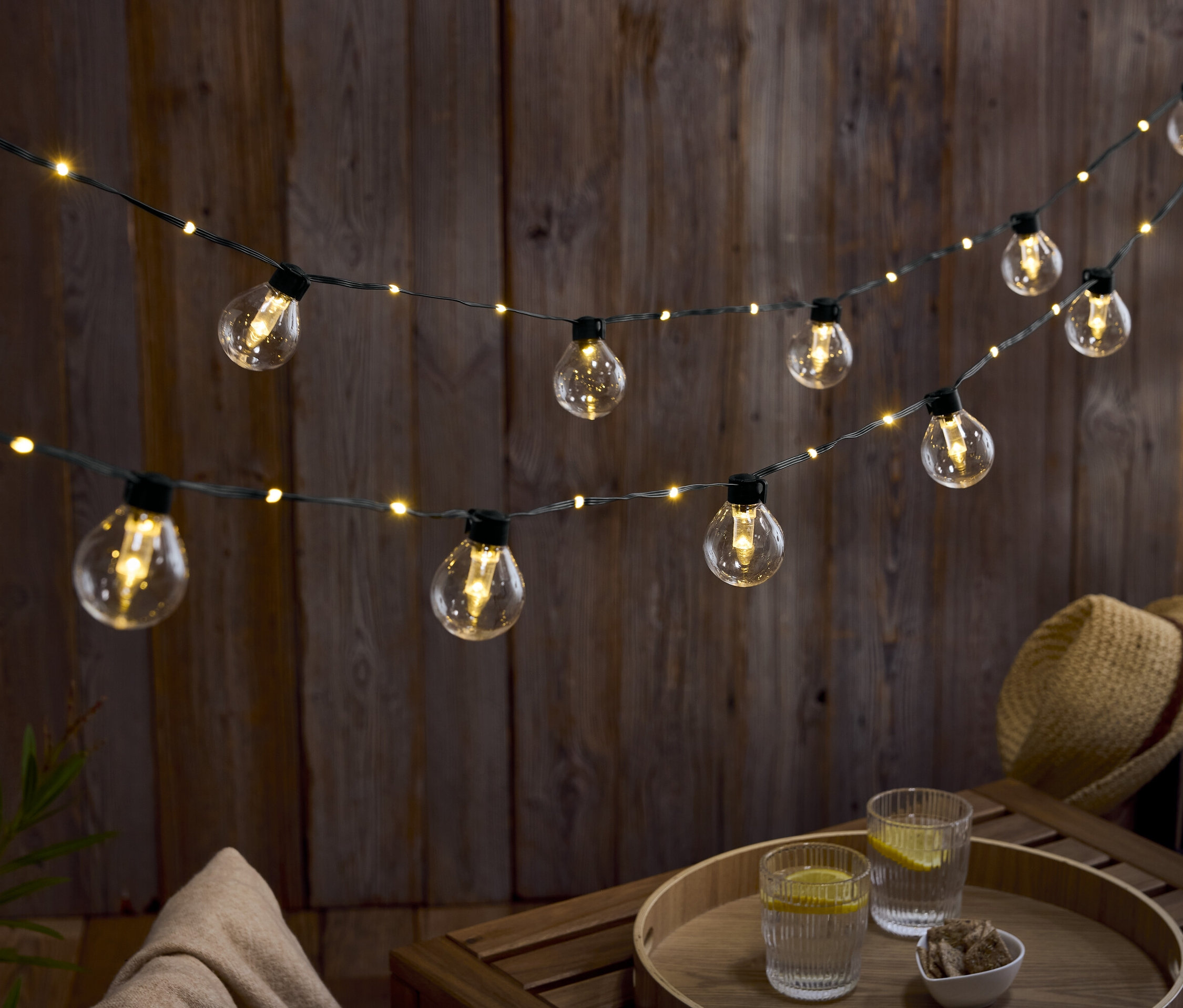 Két sor izzó lóg egy fal előtt. Egy faasztalon két pohár viz citrommal és egy tál dió.