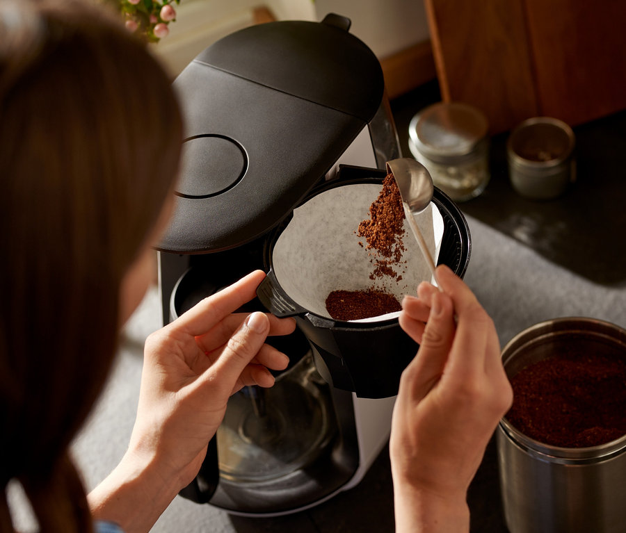 Nő kanalazza a kávét a »Let's Brew« filteres kávéfőző fehér szűrőjébe.
