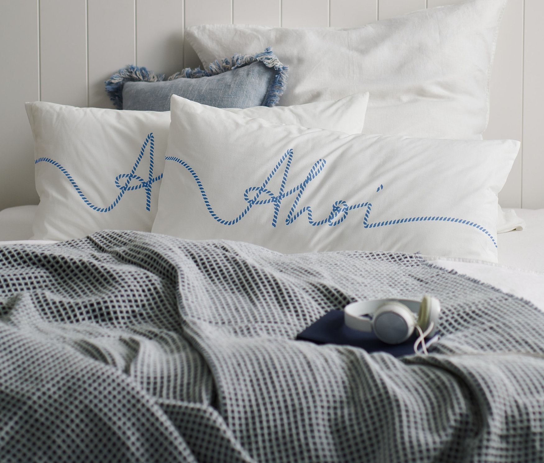 Két fehér »Ahoi« perkál párnahuzat kék felirattal egy ágyon, szürke takaróval és fejhallgatóval.