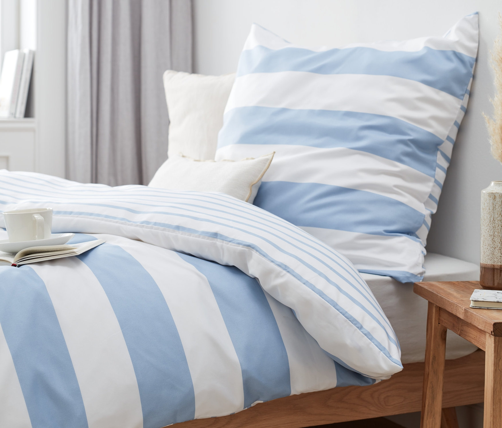 Kék csíkos mikroszálas ágynemű (normál és nagy méretű) egy ágyon, könyvvel és csészével.