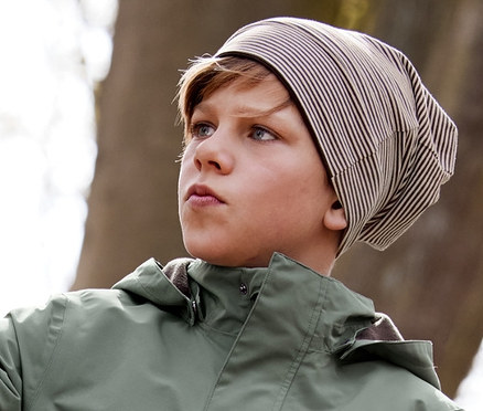 Fiú dől egy fának, barna, kifordítható sapkában, fiú kabátban minden időjárásra, fiú softshell nadrágban és barna gumicsizmában.