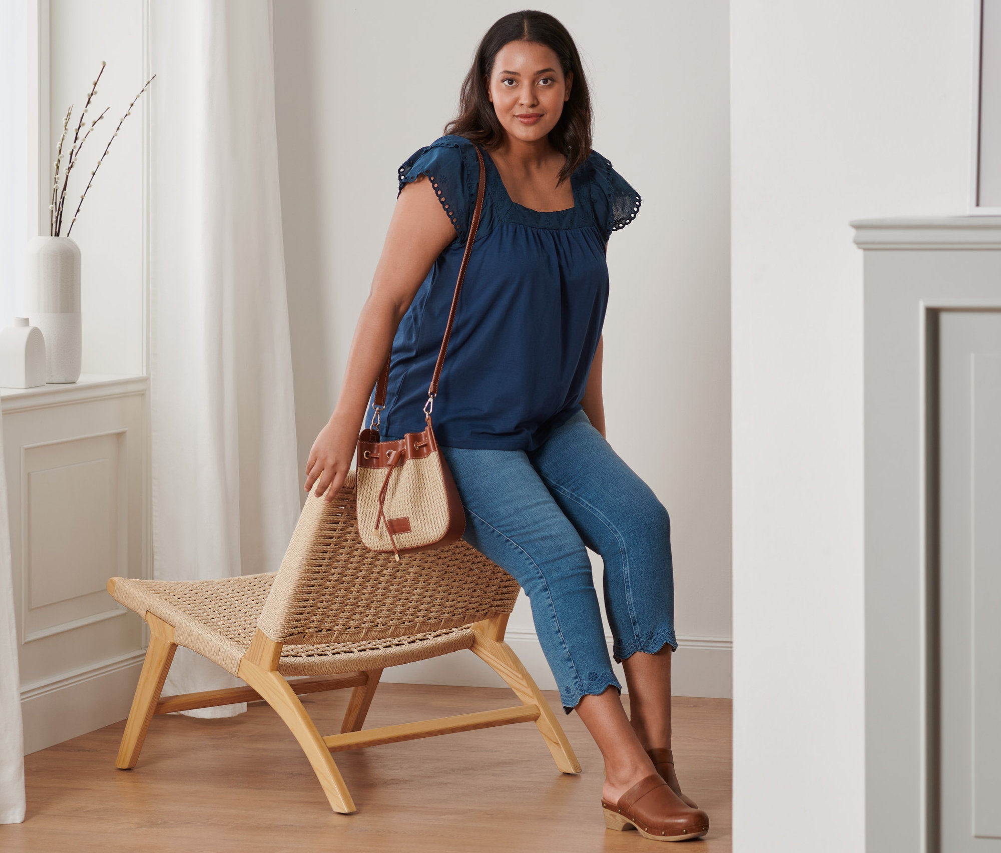 Nő ül egy fonott széken, sötétkék hímzett trikót, 7/8-os treggingst és bőr klumpát visel. Egy oldaltáska van nála.