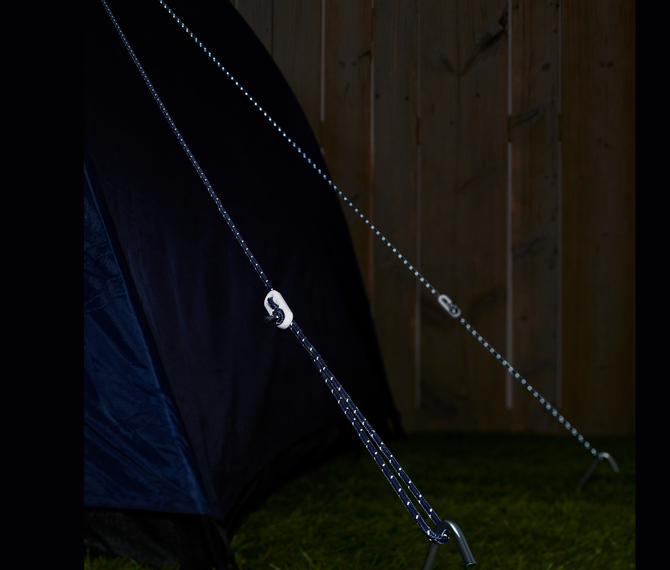 Két fényvisszaverő feszítőzsinór van rögzítve egy sátorhoz és egy cövekhez a fűben.