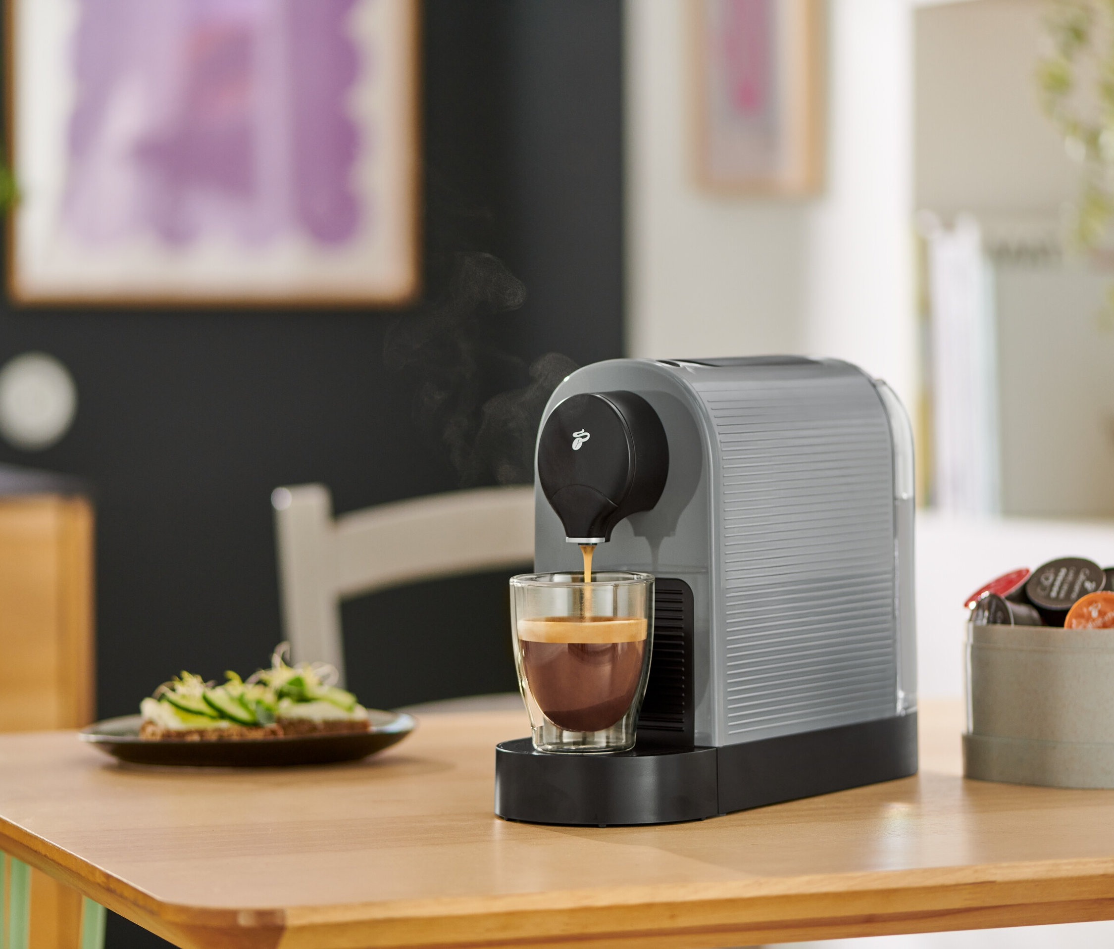 A szürke Cafissimo pure+ kávéfőző kávét tölt egy pohárba. Az asztalon egy tányér szendvics.