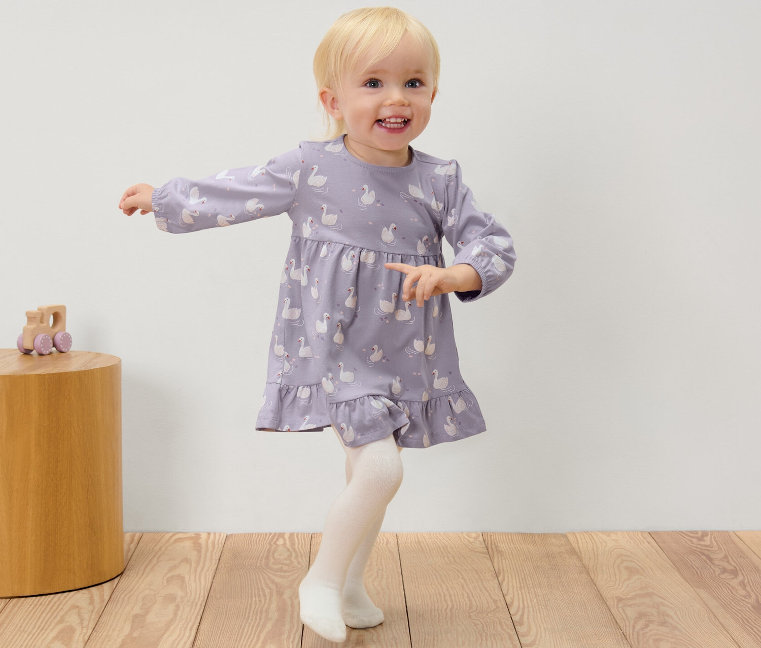 Kislány hattyúmintás ruhában és harisnyában áll egy faasztal mellett, amin egy játék van.