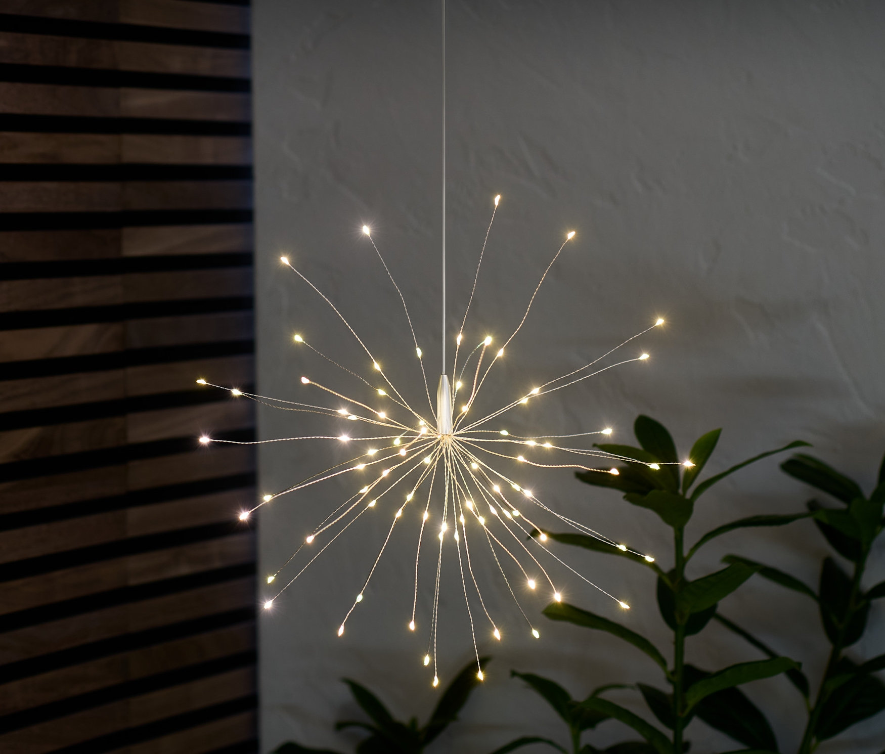 Egy LED-es világító ág lóg és világít a szobában egy növény mellett.