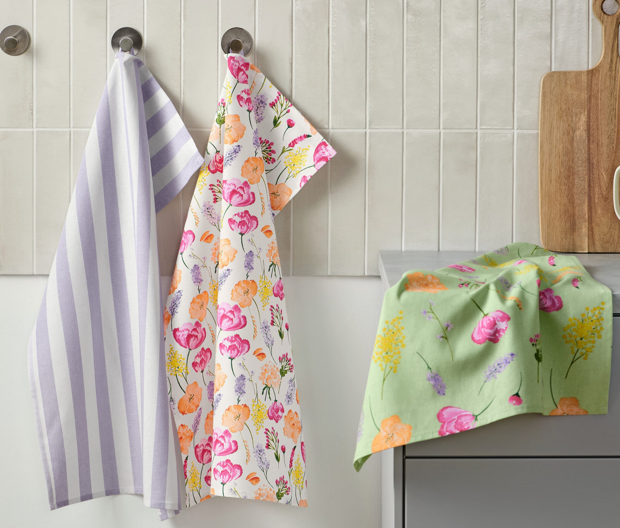 Három konyharuha lóg a falon: egy lila-fehér csíkos, egy virágmintás, egy pedig a pulton van. Mellette egy vágódeszka és egy bögre konyhai eszközökkel. 3 konyharuha, virágos/csíkos