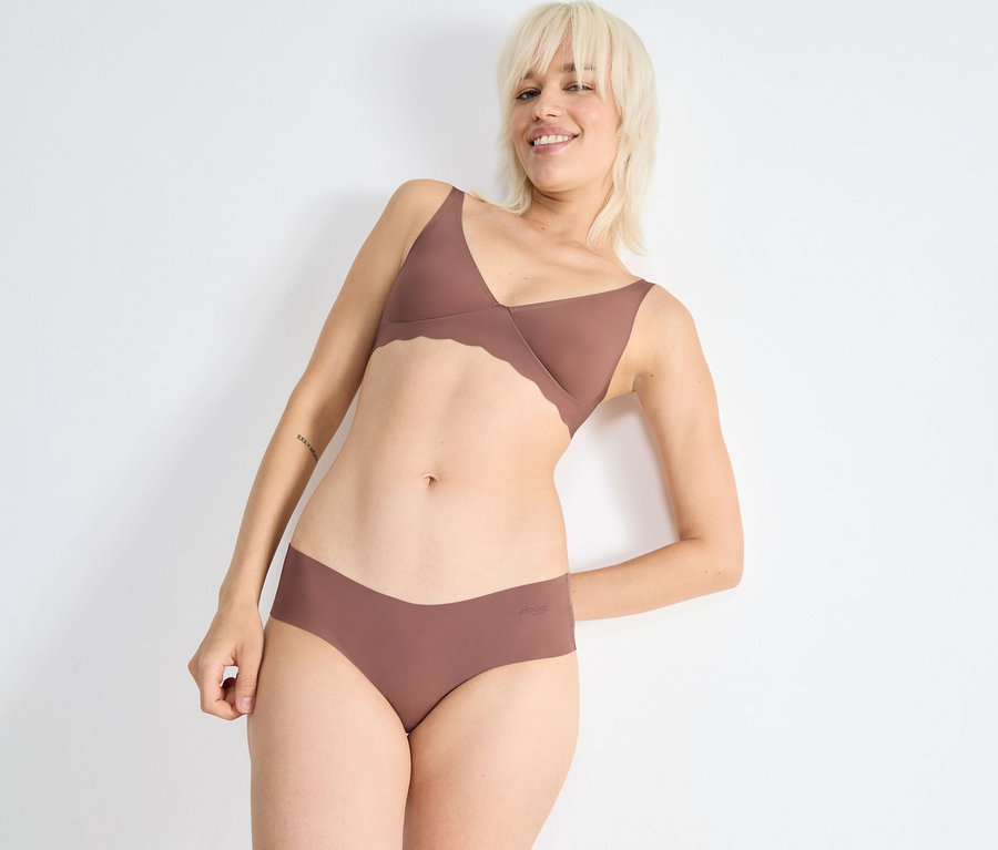 Mosolygó nő rövid szőke hajjal fehér falnak támaszkodva, barna-rózsaszín hullámos szélű melltartót és sloggi ZERO Microfibre 2.0 hipszter alsót visel.
