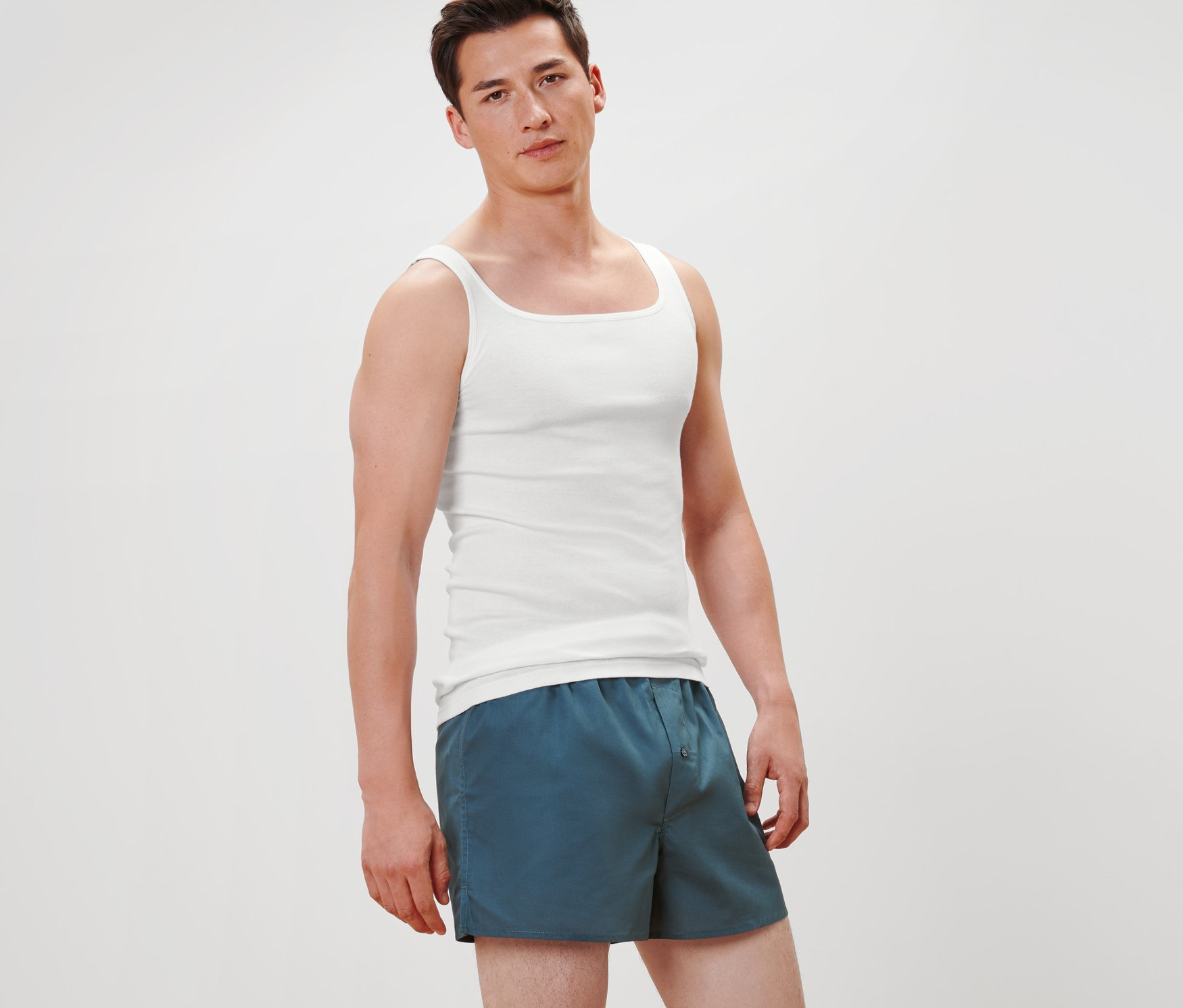 Egy férfi áll, és 2 fehér, minőségi, finoman bordázott trikót és kék rövidnadrágot visel.