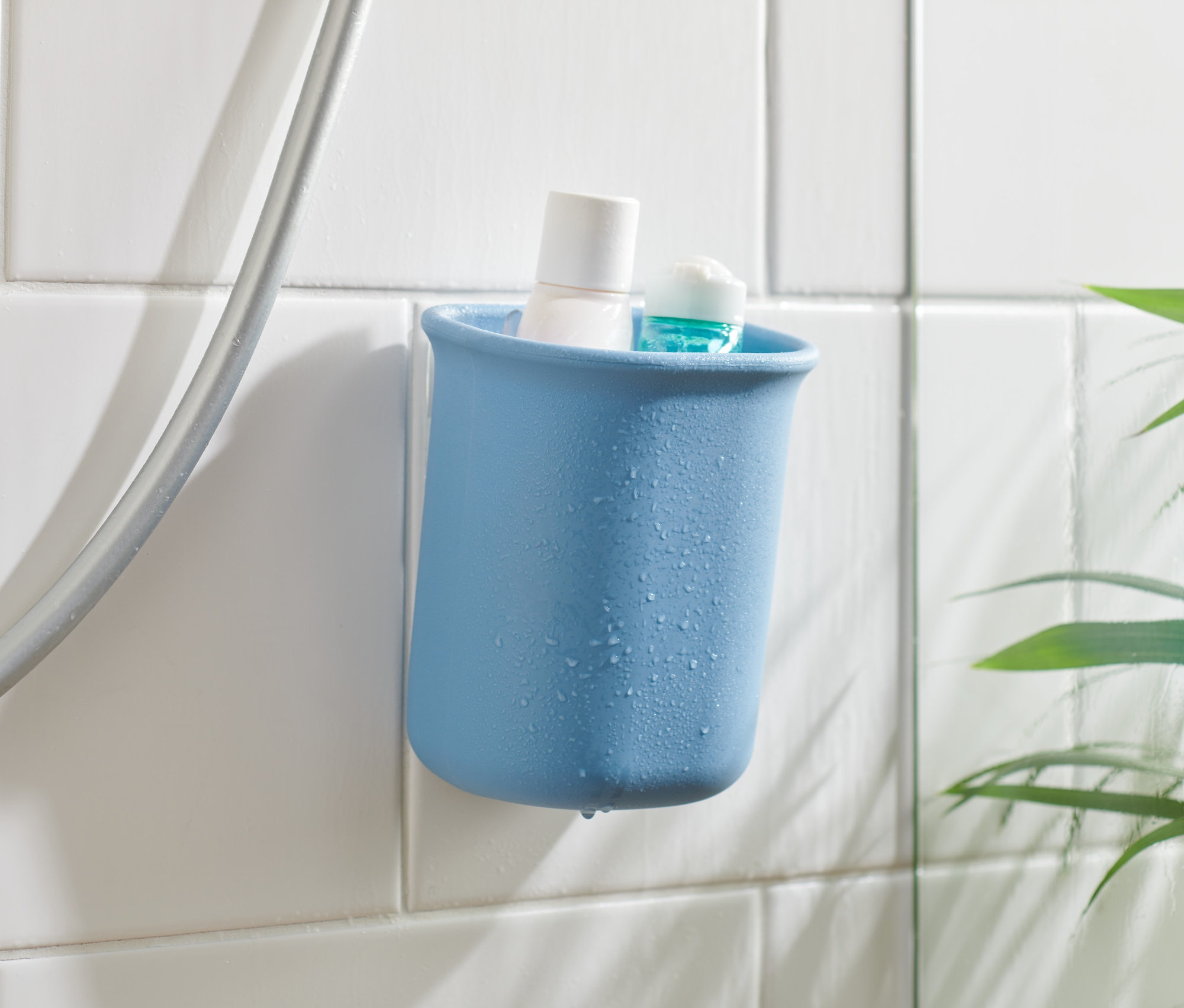 Kék falikiegészítő-tartó fehér és kék termékekkel, fehér csempére rögzítve a zuhanycső mellett.