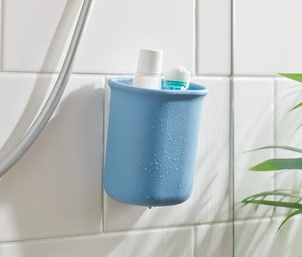 Kék falikiegészítő-tartó fehér és kék termékekkel, fehér csempére rögzítve a zuhanycső mellett.