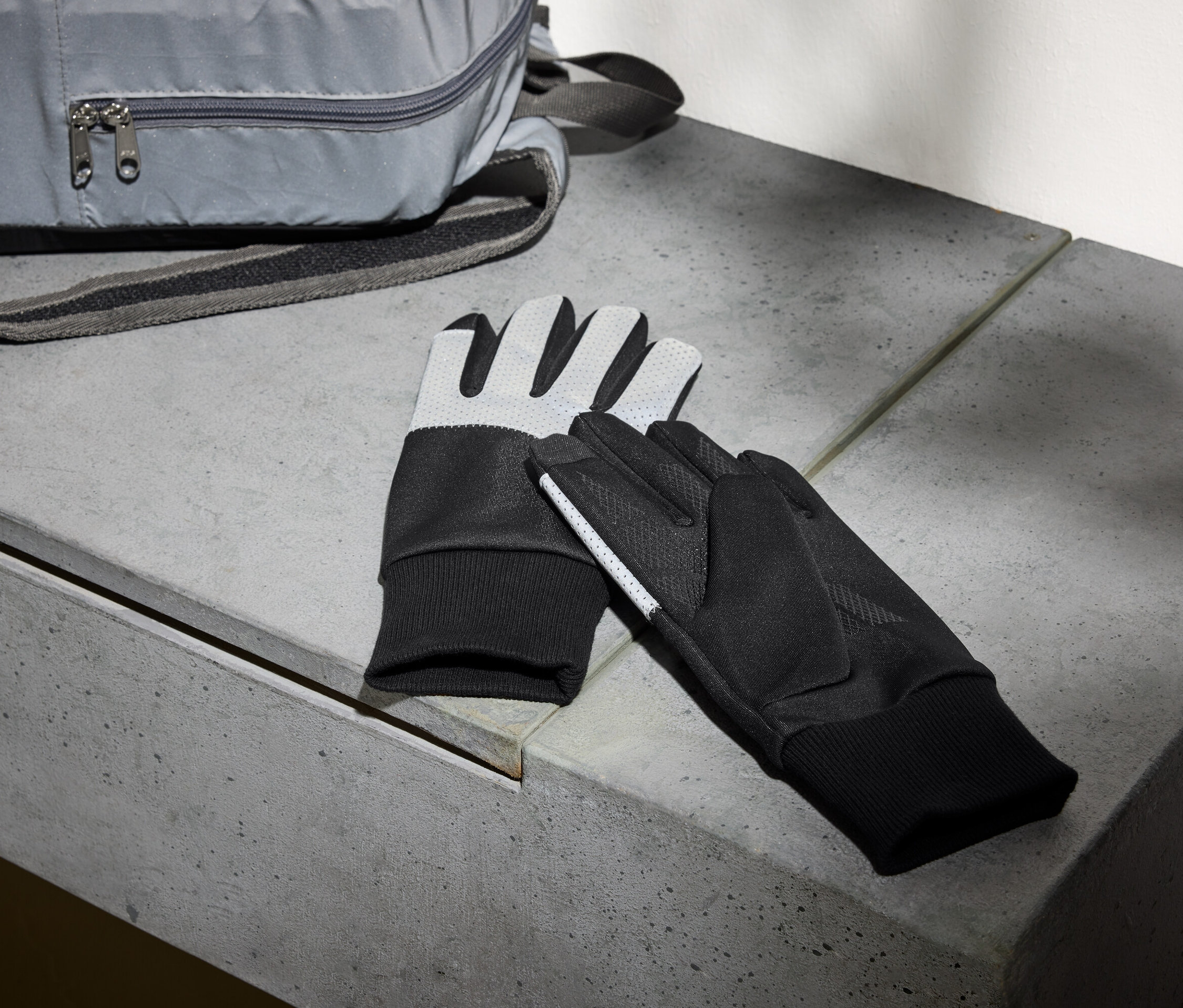 Egy szürke hátizsák és egy pár fekete kesztyű fekszik egy betonpadon.