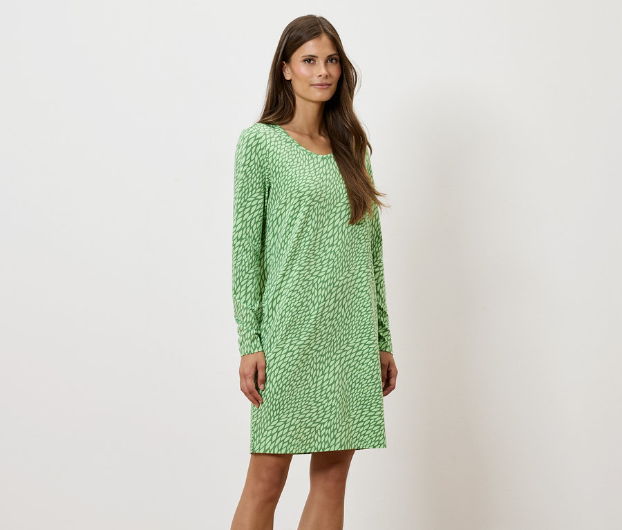 Egy nő áll és pózol egy zöld, mintás, hosszú ujjú ruhában.