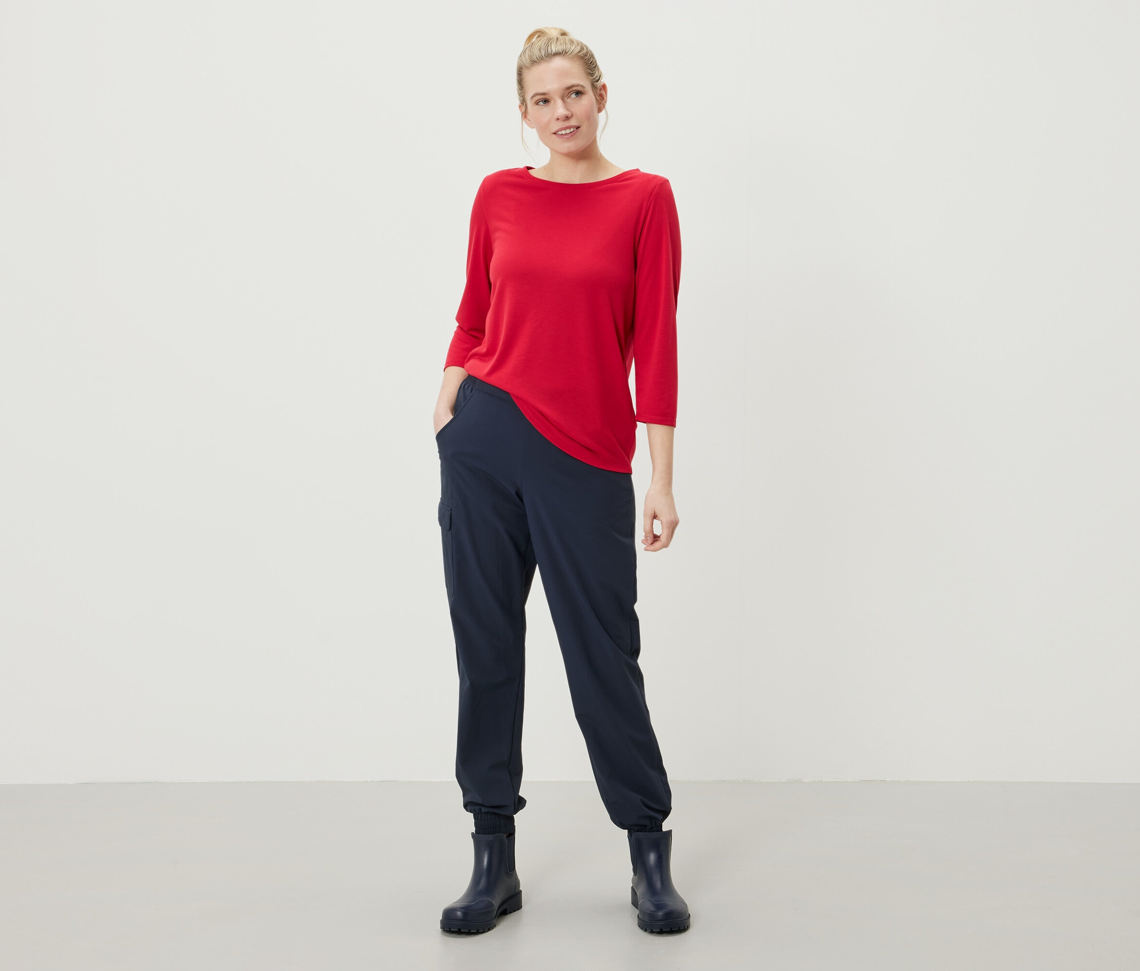 Egy nő áll, kezével a sötétkék nadrágja zsebében, piros, háromnegyedes ujjú felsőt és sötétkék gumicsizmát visel.