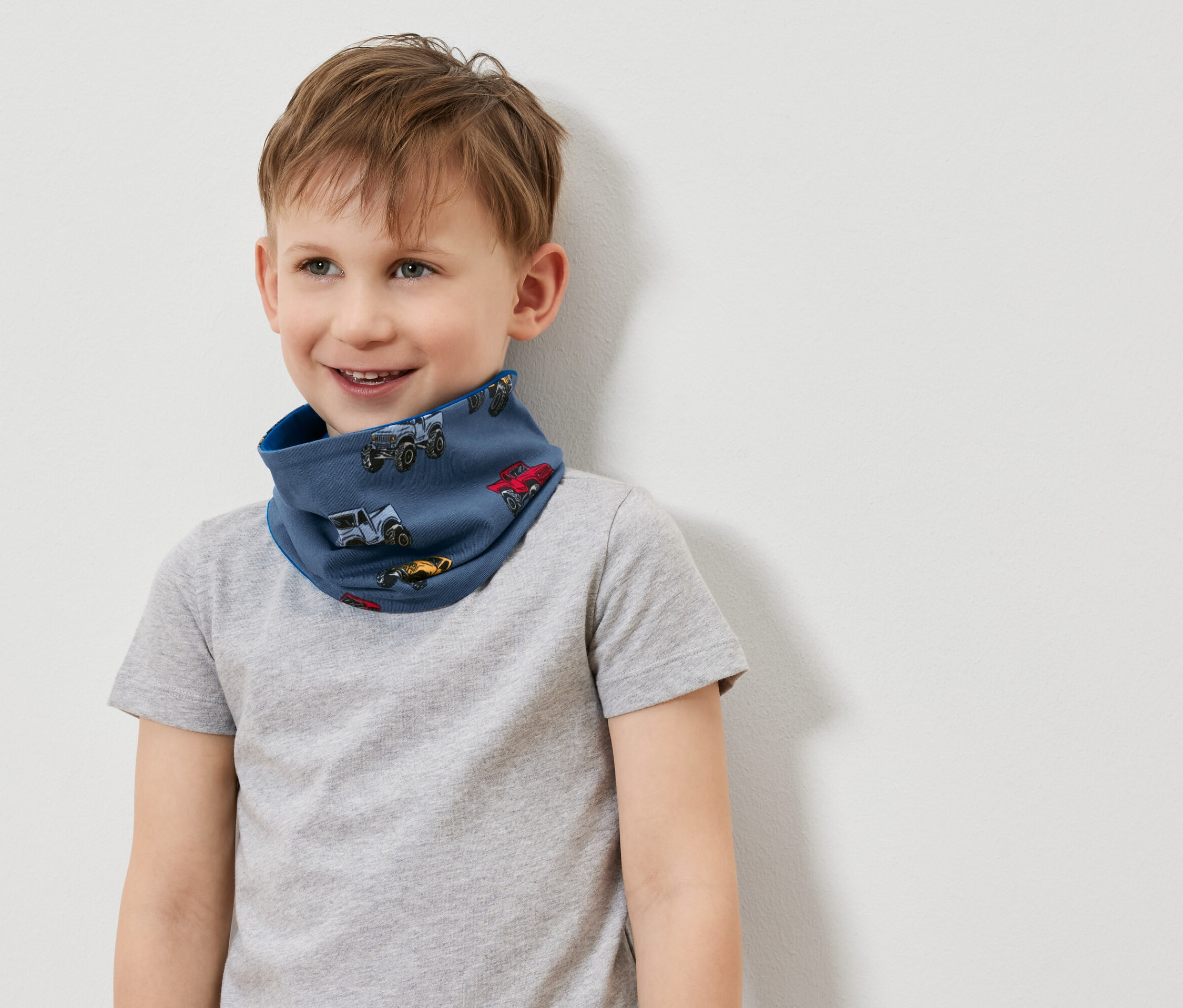 Mosolygó fiú kék, autós mintás nyakmelegítőt és szürke pólót visel.