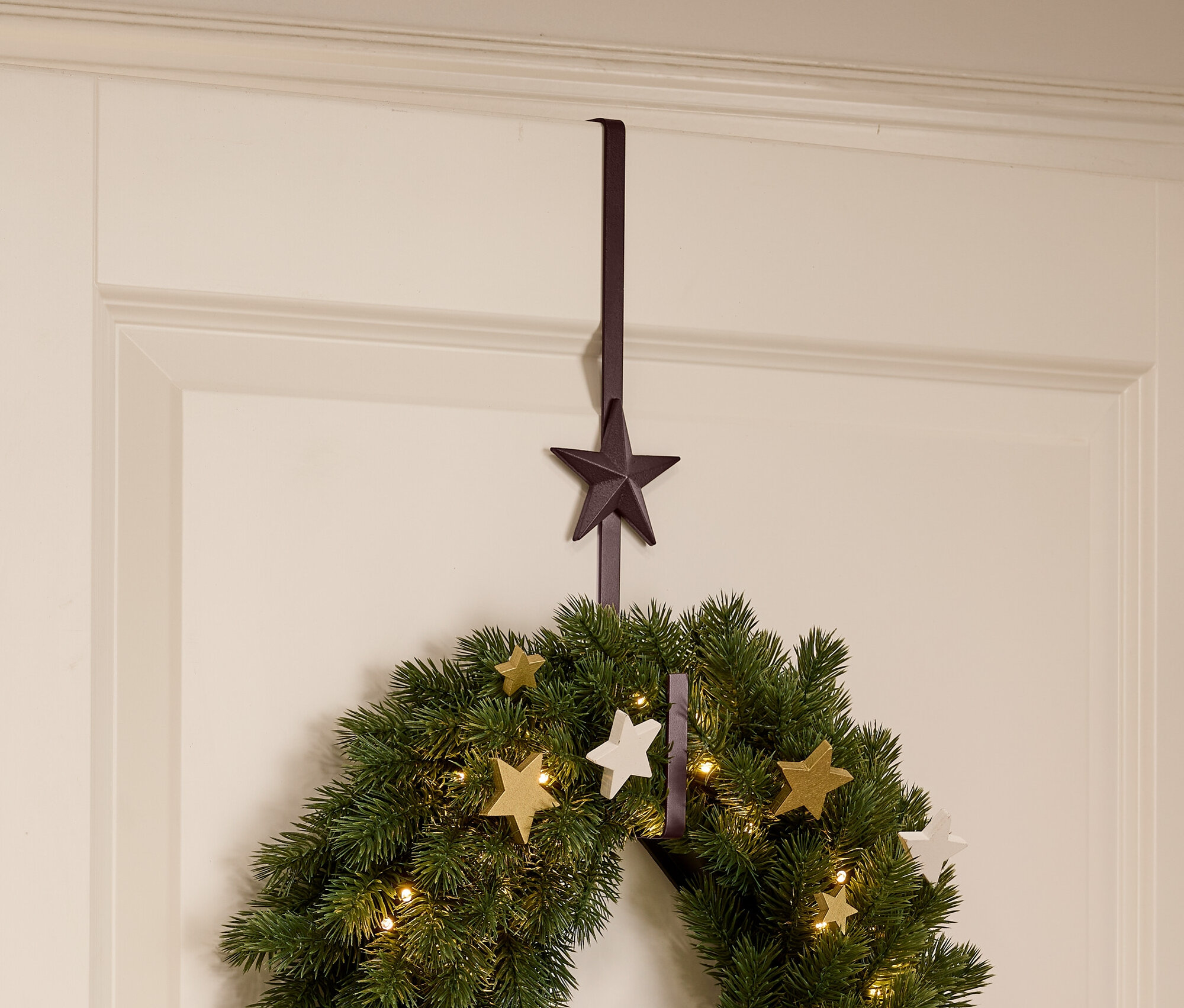 Zöld karácsonyi koszorú csillagokkal és fényekkel lóg egy fehér ajtón egy barna csillag alakú akasztóval.